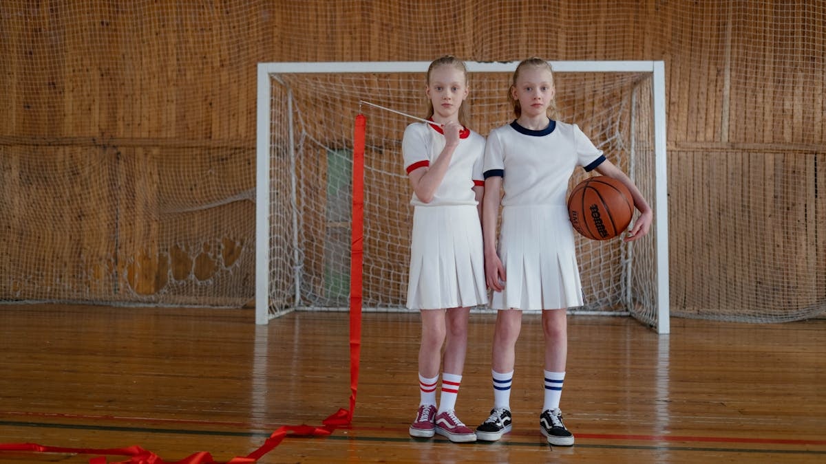 Twin girls in sports outfits stand in a gym with a basketball and rhythmic gymnastics ribbon.